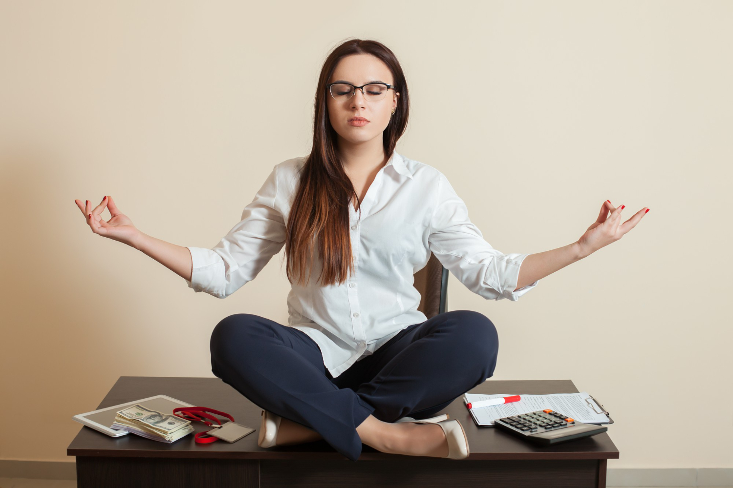 Woman in glasses sitting cross-legged on an office desk with eyes closed, practising calm and stress relief
