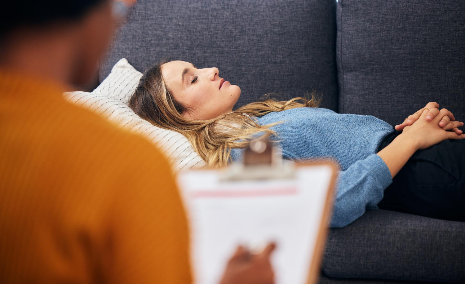 Hypnotherapist with a clipboard beside a relaxed client resting on a sofa during a session